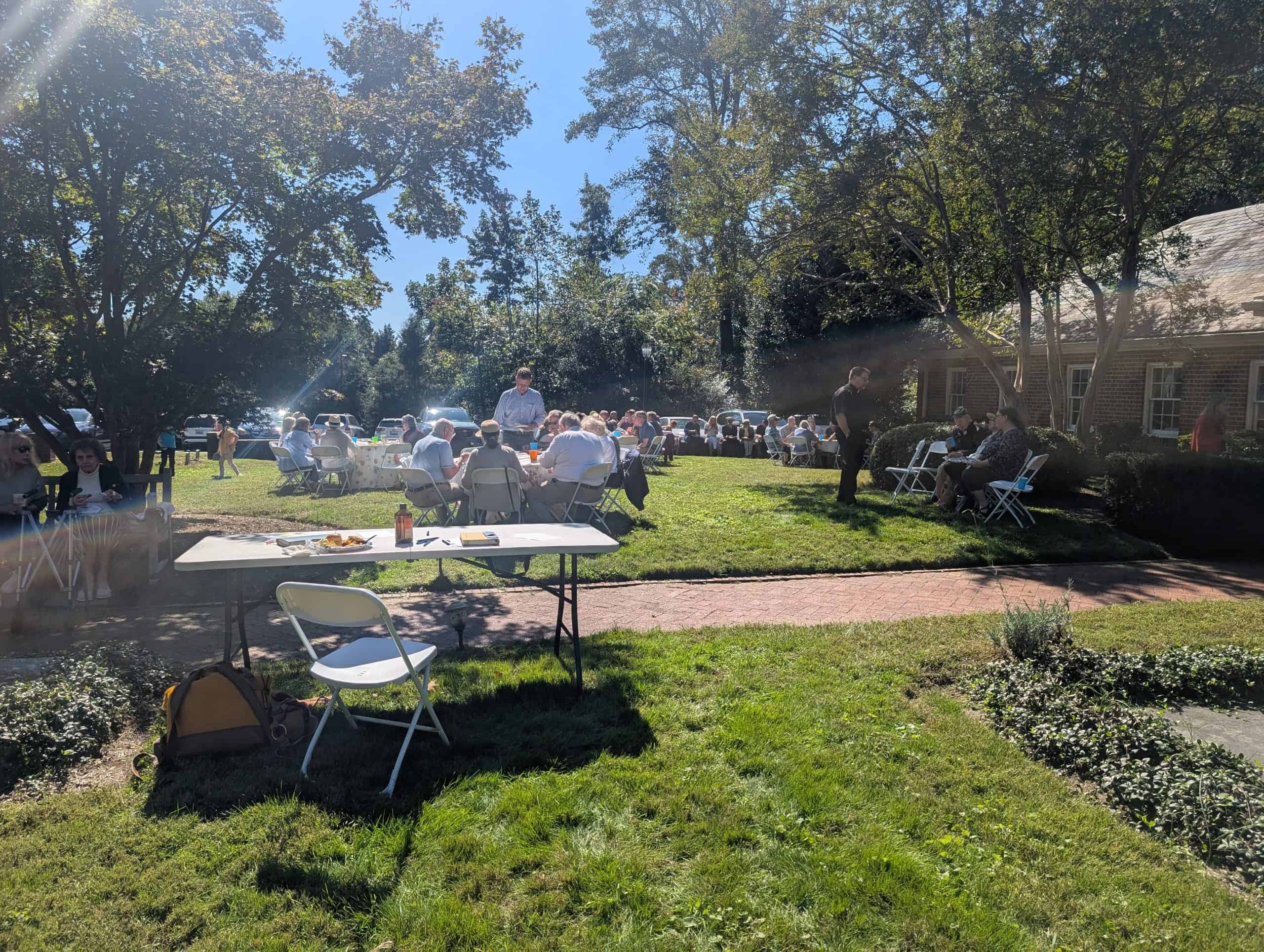 All Hallows Parish outdoor gathering with people under trees on sunny day.